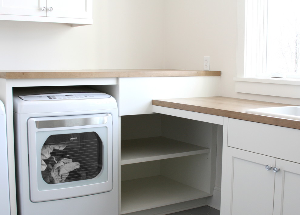 Diseño de cuarto de lavado en L campestre con armarios con paneles empotrados, puertas de armario blancas, encimera de madera, paredes blancas, lavadora y secadora juntas y suelo gris