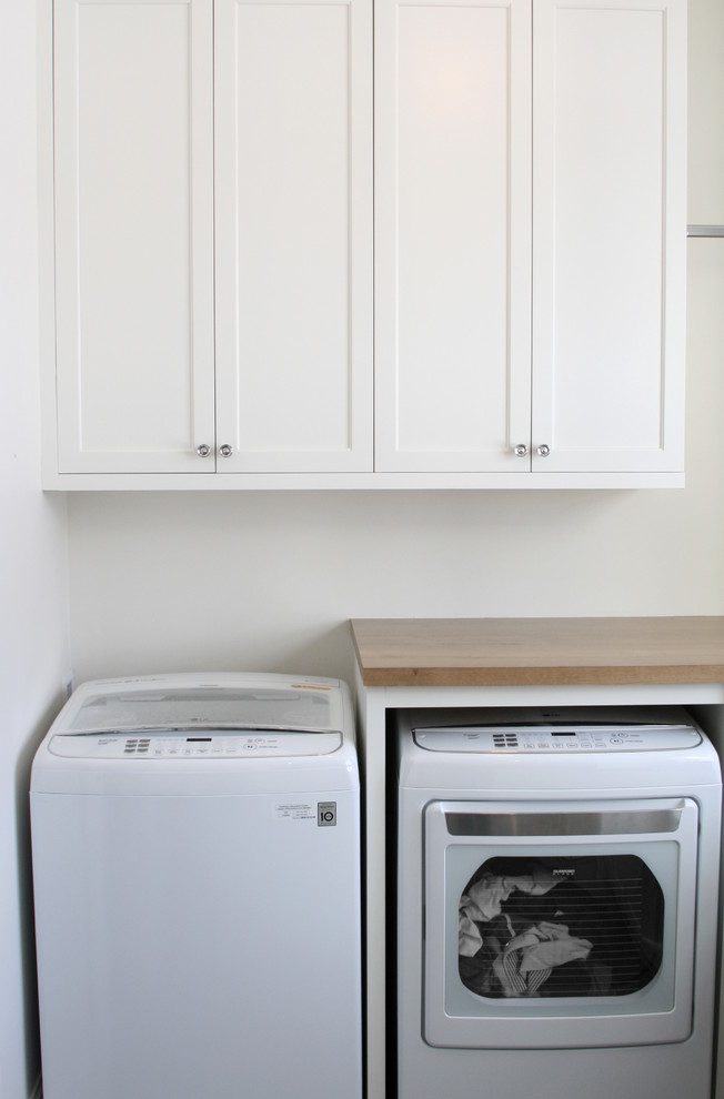 Modelo de cuarto de lavado en L campestre con armarios con paneles empotrados, puertas de armario blancas, encimera de madera, paredes blancas, lavadora y secadora juntas y suelo gris