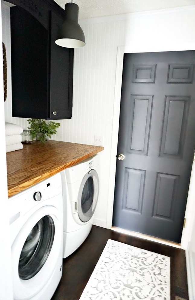 Diseño de lavadero de galera de estilo de casa de campo pequeño con puertas de armario negras, encimera de madera, paredes blancas, suelo laminado y lavadora y secadora juntas