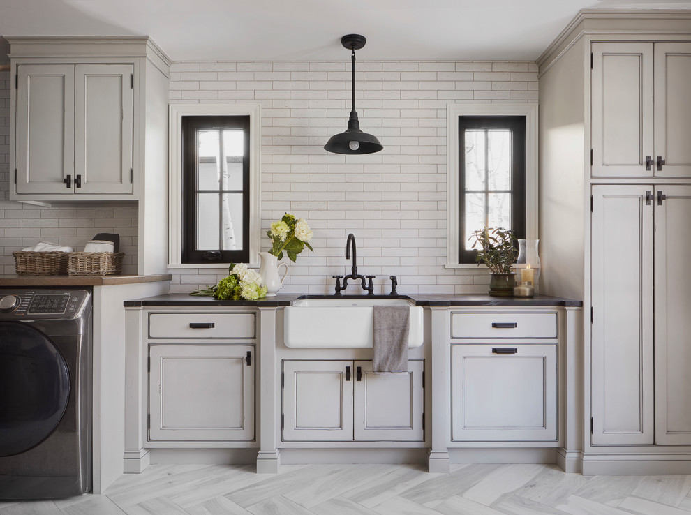 Dedicated laundry room - large cottage single-wall porcelain tile and gray floor dedicated laundry room idea in Denver with a farmhouse sink, recessed-panel cabinets, gray cabinets, soapstone countertops, white walls, a side-by-side washer/dryer and black countertops
