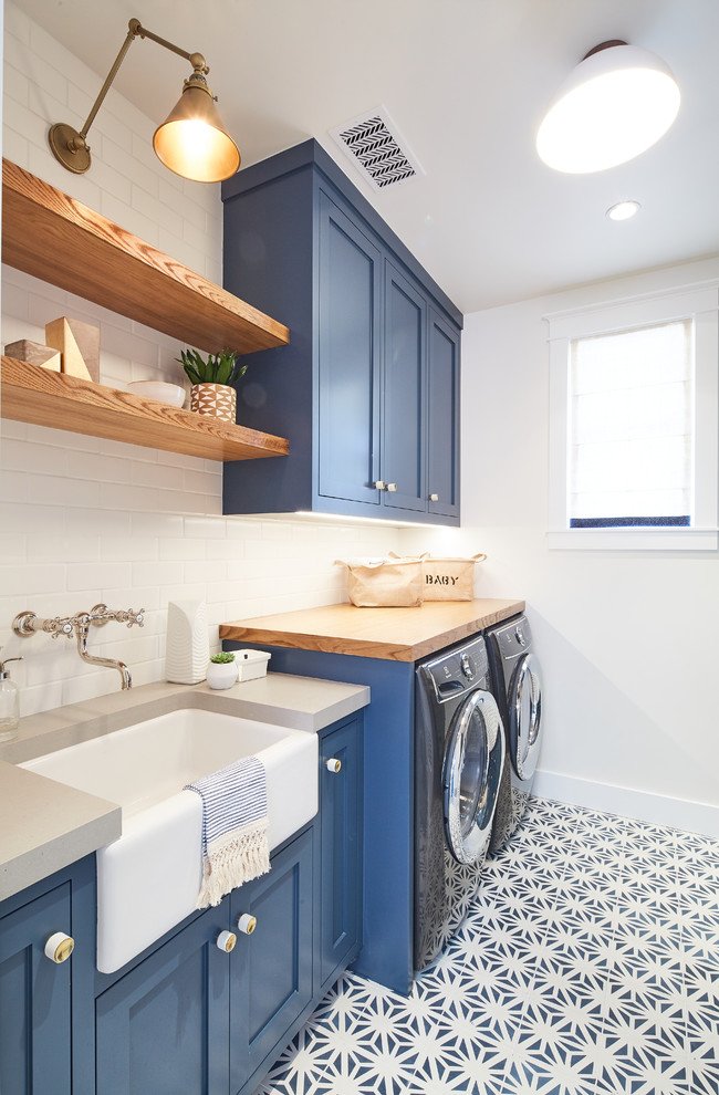Maritim inredning av en grå linjär grått tvättstuga enbart för tvätt, med en rustik diskho, skåp i shakerstil, blå skåp, träbänkskiva, vita väggar, en tvättmaskin och torktumlare bredvid varandra och flerfärgat golv