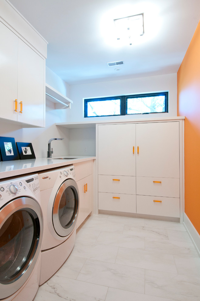 Idéer för mellanstora funkis l-formade vitt tvättstugor enbart för tvätt, med släta luckor, vita skåp, en tvättmaskin och torktumlare bredvid varandra, en undermonterad diskho, bänkskiva i kvarts, orange väggar och klinkergolv i porslin