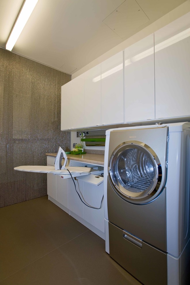 Inredning av ett modernt mellanstort parallellt grovkök, med släta luckor, vita skåp, en tvättmaskin och torktumlare bredvid varandra och grå väggar