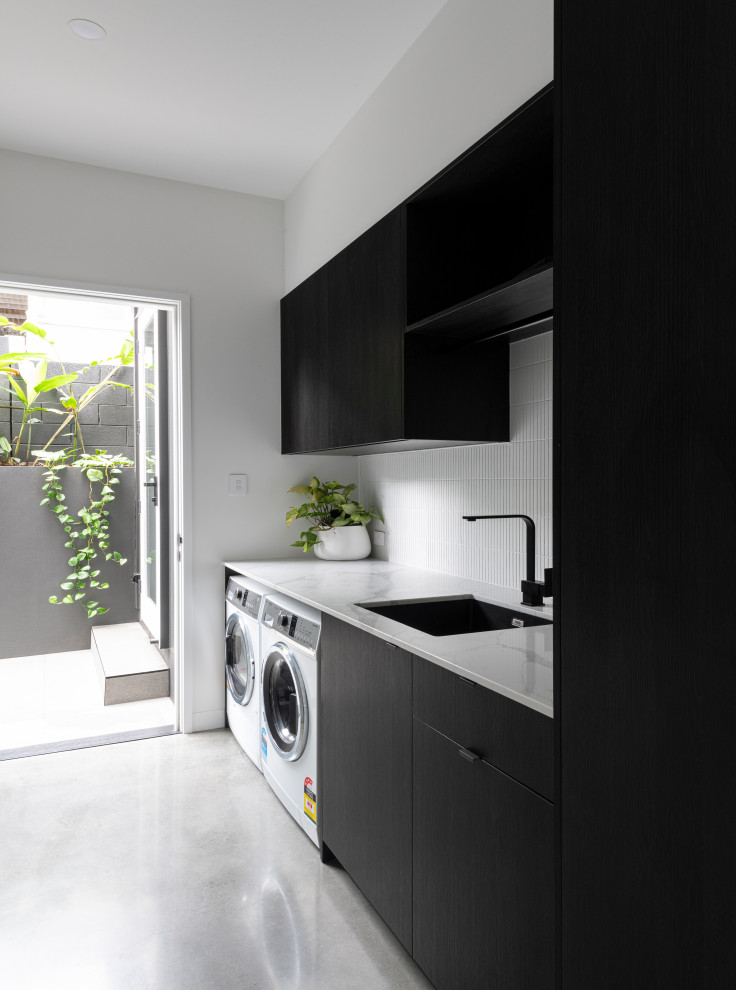 Einzeiliger Moderner Hauswirtschaftsraum mit Unterbauwaschbecken, flächenbündigen Schrankfronten, schwarzen Schränken, weißer Wandfarbe, Betonboden, Waschmaschine und Trockner nebeneinander, grauem Boden und weißer Arbeitsplatte in Brisbane