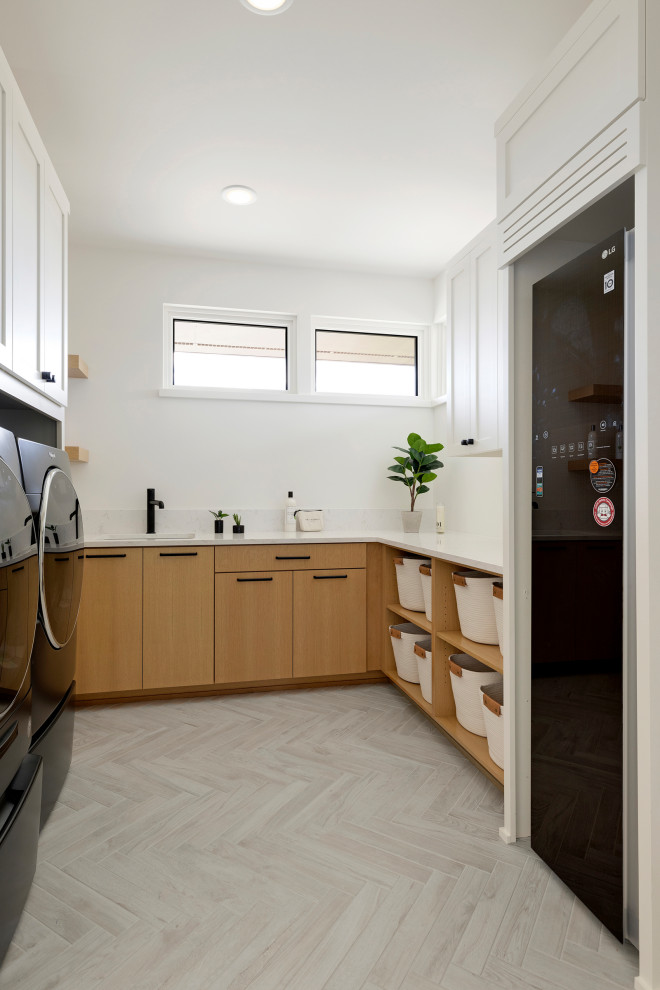 ミネアポリスにある広いカントリー風のおしゃれな洗濯室 (コの字型、アンダーカウンターシンク、フラットパネル扉のキャビネット、淡色木目調キャビネット、クオーツストーンカウンター、白いキッチンパネル、クオーツストーンのキッチンパネル、白い壁、セラミックタイルの床、左右配置の洗濯機・乾燥機、グレーの床、白いキッチンカウンター、三角天井) の写真