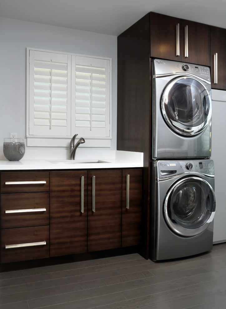 Example of a large minimalist single-wall dark wood floor dedicated laundry room design in San Francisco with an undermount sink, flat-panel cabinets, distressed cabinets, quartz countertops, gray walls, a stacked washer/dryer and white countertops