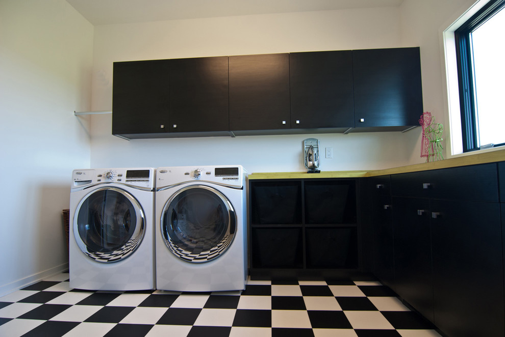 グランドラピッズにある低価格の中くらいなモダンスタイルのおしゃれな洗濯室 (L型、フラットパネル扉のキャビネット、黒いキャビネット、ラミネートカウンター、白い壁、セラミックタイルの床、左右配置の洗濯機・乾燥機、マルチカラーの床、緑のキッチンカウンター) の写真