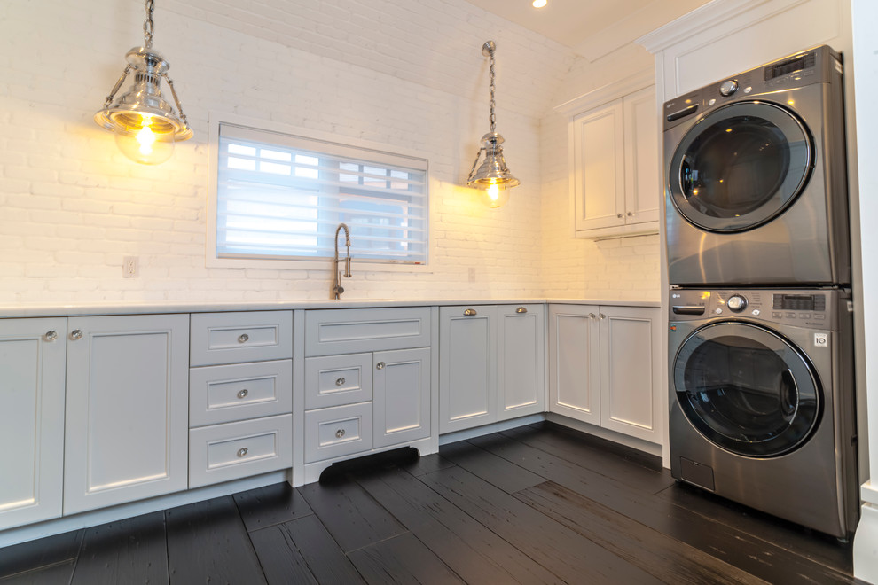 Diseño de cuarto de lavado en L clásico grande con armarios estilo shaker, puertas de armario blancas, encimera de mármol, paredes blancas, suelo de madera oscura y lavadora y secadora apiladas