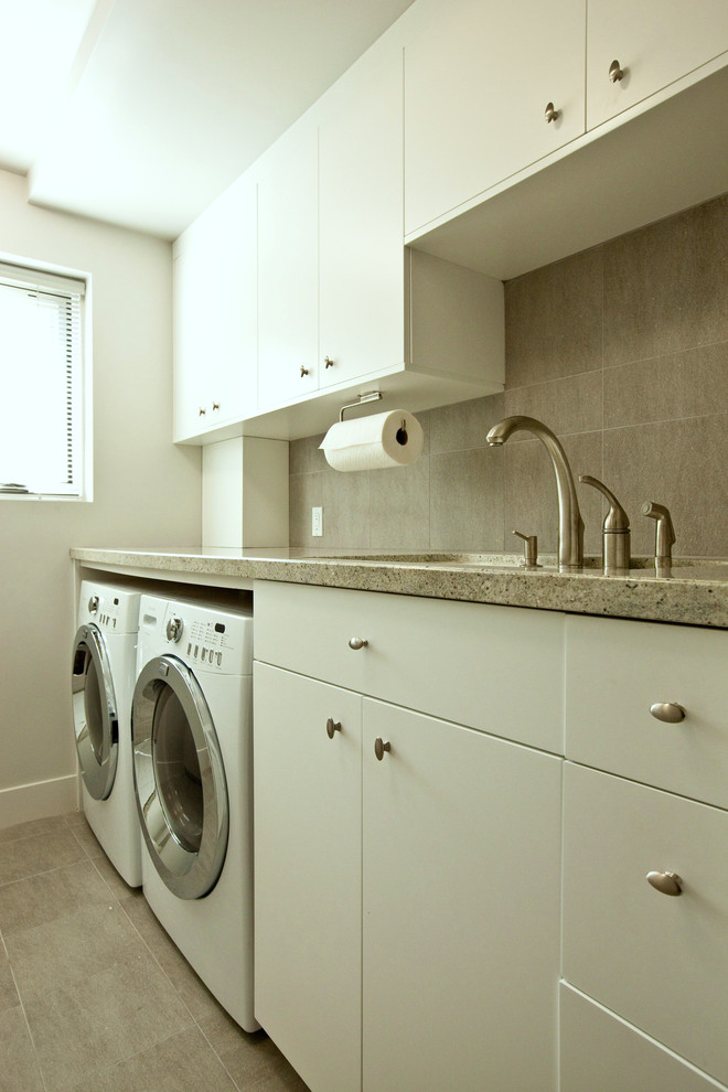 Inspiration för en stor funkis parallell tvättstuga enbart för tvätt, med en undermonterad diskho, släta luckor, vita skåp, granitbänkskiva, grå väggar, klinkergolv i porslin, en tvättmaskin och torktumlare bredvid varandra och grått golv
