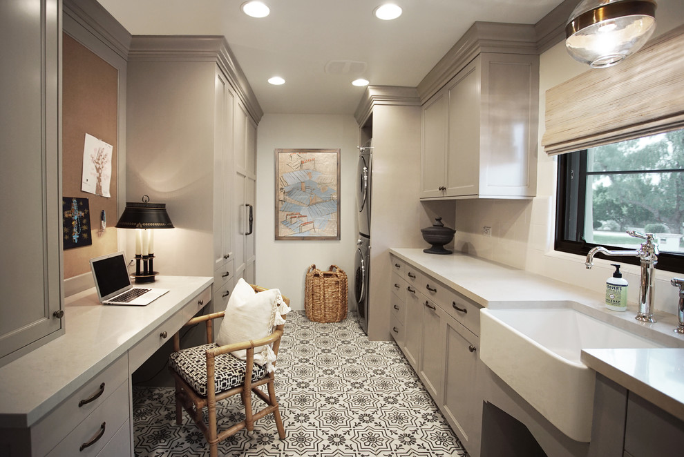 Mid-sized transitional galley multicolored floor dedicated laundry room photo in Phoenix with a farmhouse sink, recessed-panel cabinets, gray cabinets, quartz countertops, white backsplash, subway tile backsplash, white walls, a stacked washer/dryer and gray countertops