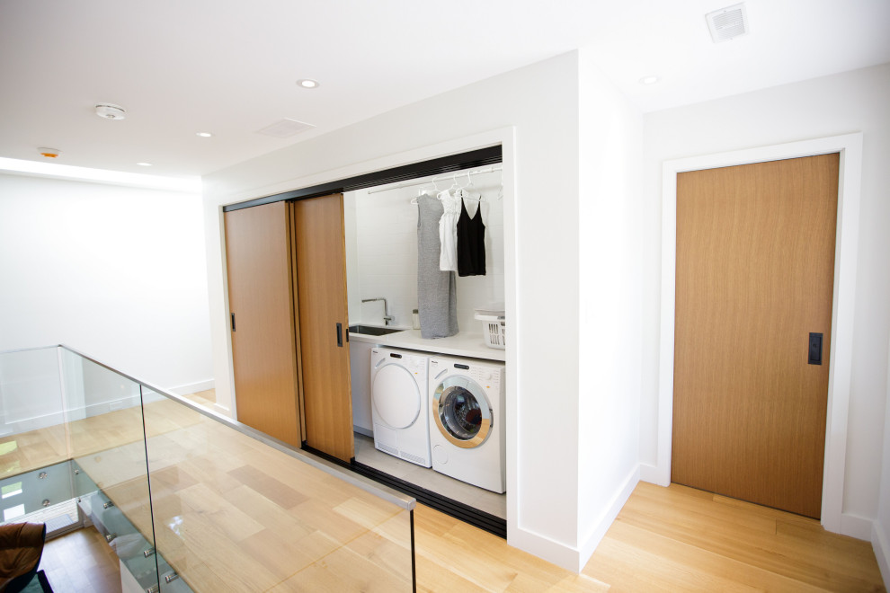 Moderner Hauswirtschaftsraum mit Doppelwaschbecken, weißen Schränken, weißer Wandfarbe, braunem Holzboden und Waschmaschine und Trockner gestapelt in Toronto