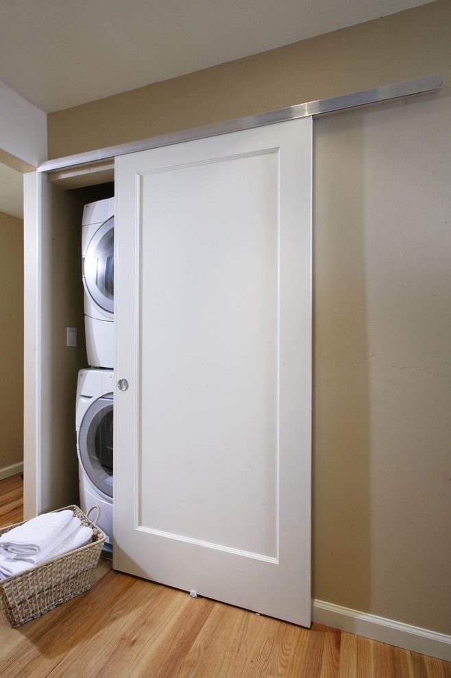 Modelo de armario lavadero lineal minimalista pequeño con suelo de madera clara, lavadora y secadora apiladas y paredes beige