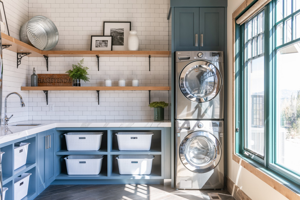 Bild på en stor rustik vita l-formad vitt tvättstuga enbart för tvätt, med en undermonterad diskho, öppna hyllor, blå skåp, vita väggar, en tvättpelare och grått golv