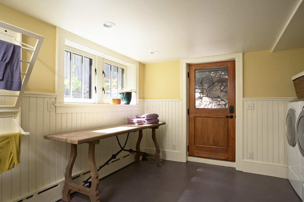 Exempel på en liten amerikansk tvättstuga, med gula väggar, en tvättmaskin och torktumlare bredvid varandra, vita skåp, träbänkskiva och betonggolv