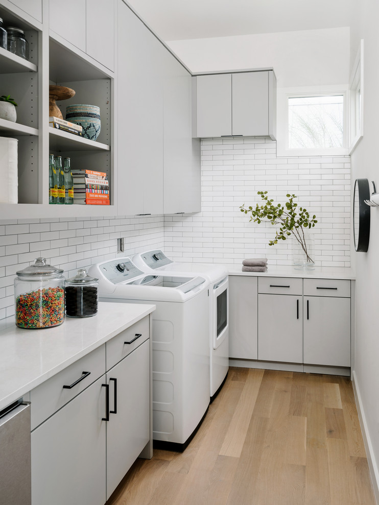 Moderne Waschküche in L-Form mit flächenbündigen Schrankfronten, grauen Schränken, weißer Wandfarbe, braunem Holzboden, Waschmaschine und Trockner nebeneinander, braunem Boden und weißer Arbeitsplatte in Austin