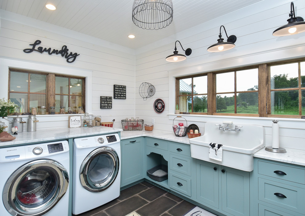 Réalisation d'une buanderie champêtre en U multi-usage avec un évier de ferme, un placard à porte shaker, des portes de placard bleues, plan de travail en marbre, un mur blanc, un sol en carrelage de porcelaine, un lave-linge séchant, un sol noir et un plan de travail blanc.