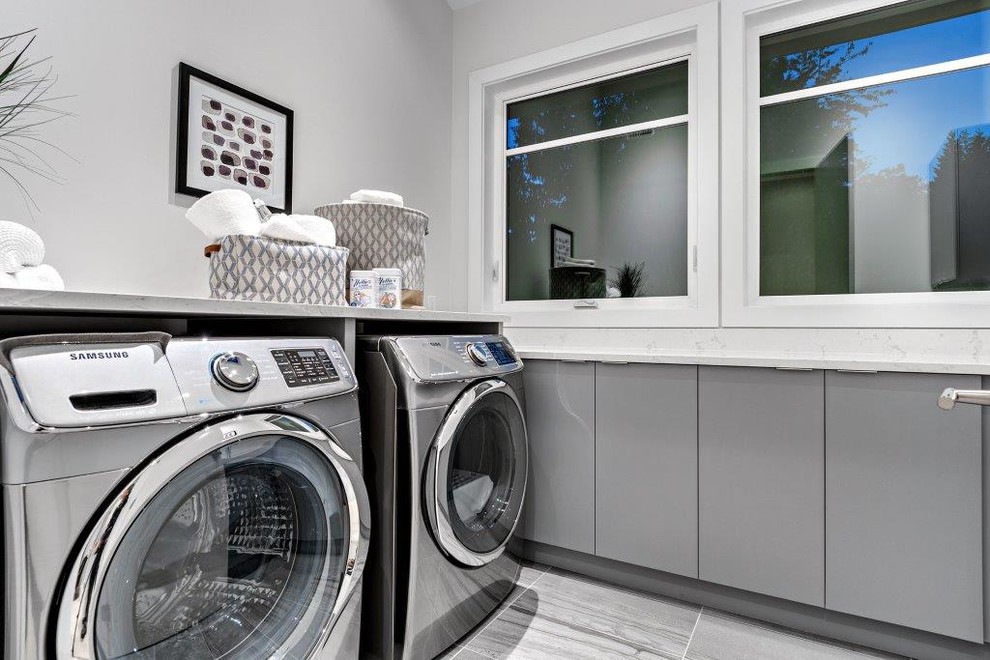 Modern inredning av en u-formad tvättstuga enbart för tvätt, med en undermonterad diskho, släta luckor, grå skåp, bänkskiva i kvarts, grå väggar, klinkergolv i porslin och en tvättmaskin och torktumlare bredvid varandra
