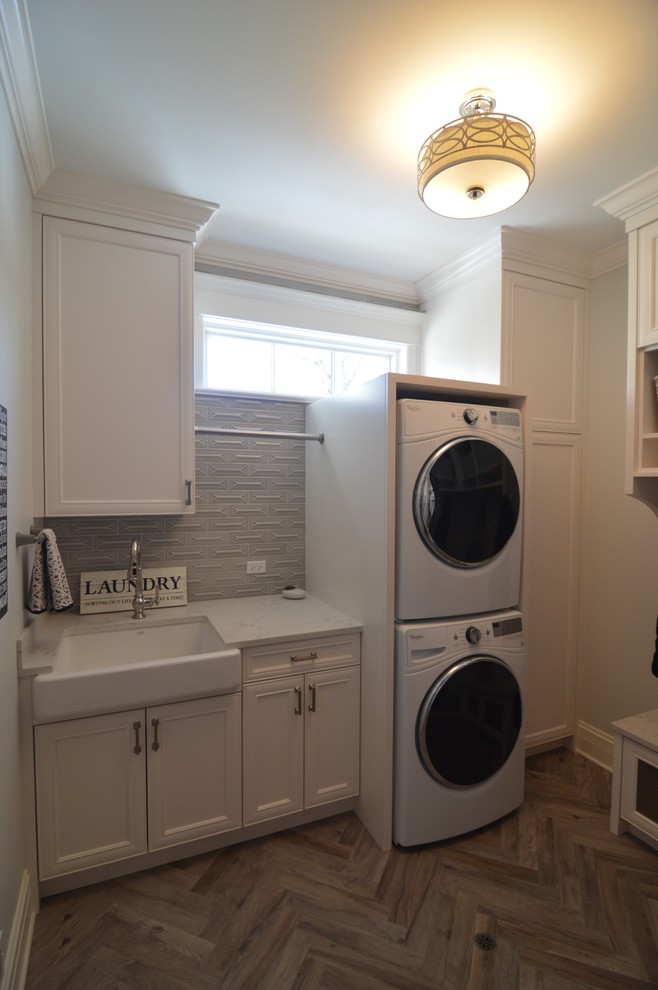 Photo of a medium sized classic utility room in Chicago with a belfast sink, recessed-panel cabinets, white cabinets, engineered stone countertops, grey walls, porcelain flooring, a stacked washer and dryer and brown floors.