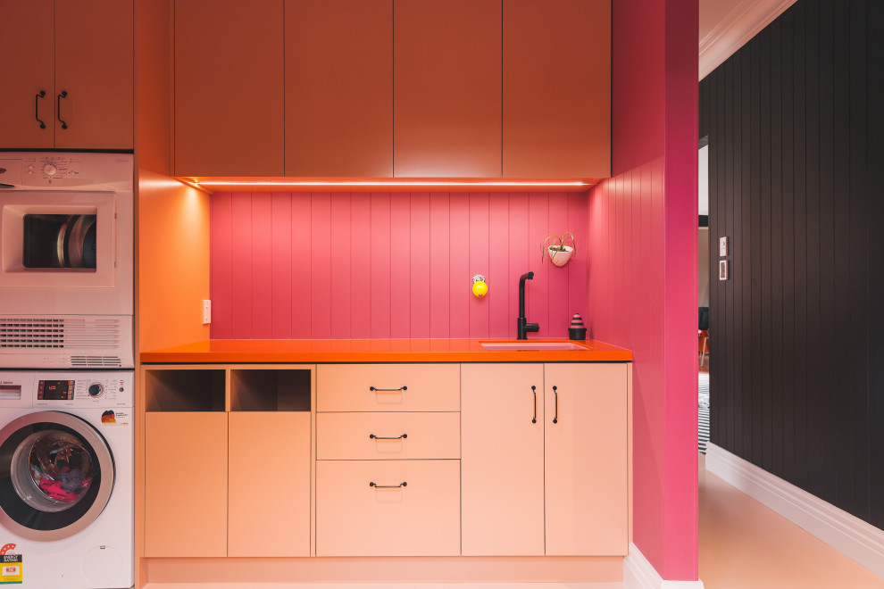 他の地域にあるエクレクティックスタイルのおしゃれなランドリールーム (ll型、アンダーカウンターシンク、レイズドパネル扉のキャビネット、オレンジのキャビネット、ラミネートカウンター、ピンクのキッチンパネル、木材のキッチンパネル、ピンクの壁、塗装フローリング、上下配置の洗濯機・乾燥機、ピンクの床、オレンジのキッチンカウンター、塗装板張りの天井、羽目板の壁) の写真