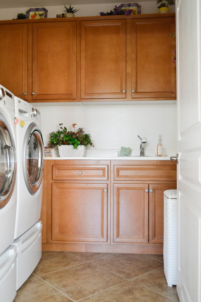 Kleine Mediterrane Waschküche in L-Form mit Waschbecken, hellbraunen Holzschränken, weißer Wandfarbe, Travertin und Waschmaschine und Trockner nebeneinander in Los Angeles