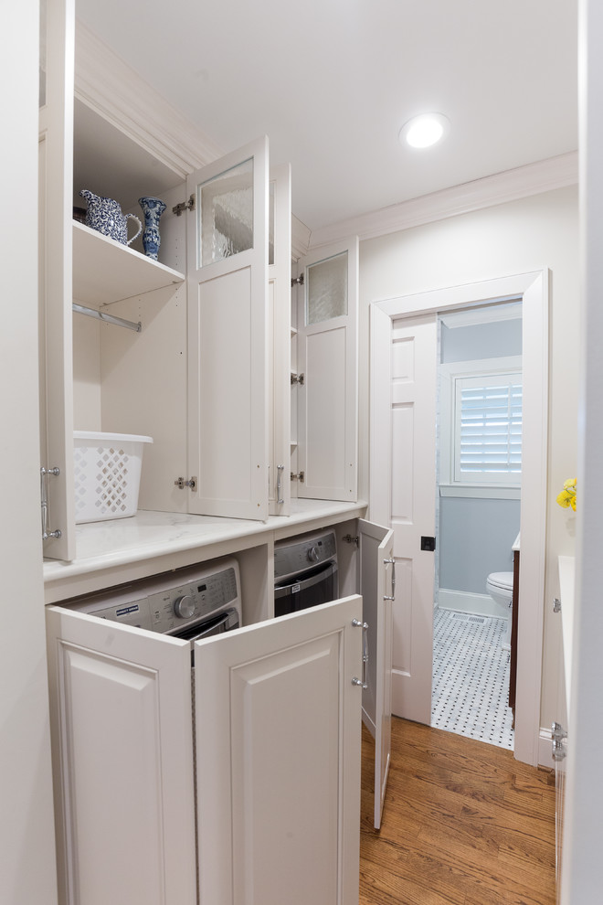 This is an example of a small transitional single-wall laundry cupboard in Charlotte with raised-panel cabinets, white cabinets, quartzite benchtops, white walls, medium hardwood floors, a concealed washer and dryer and brown floor.