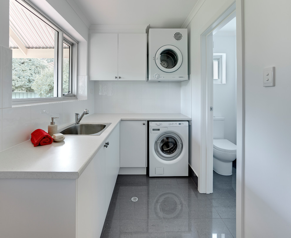 Inspiration for a mid-sized contemporary l-shaped porcelain tile and gray floor dedicated laundry room remodel in Other with a single-bowl sink, white cabinets, laminate countertops, white walls, a stacked washer/dryer, flat-panel cabinets and white countertops