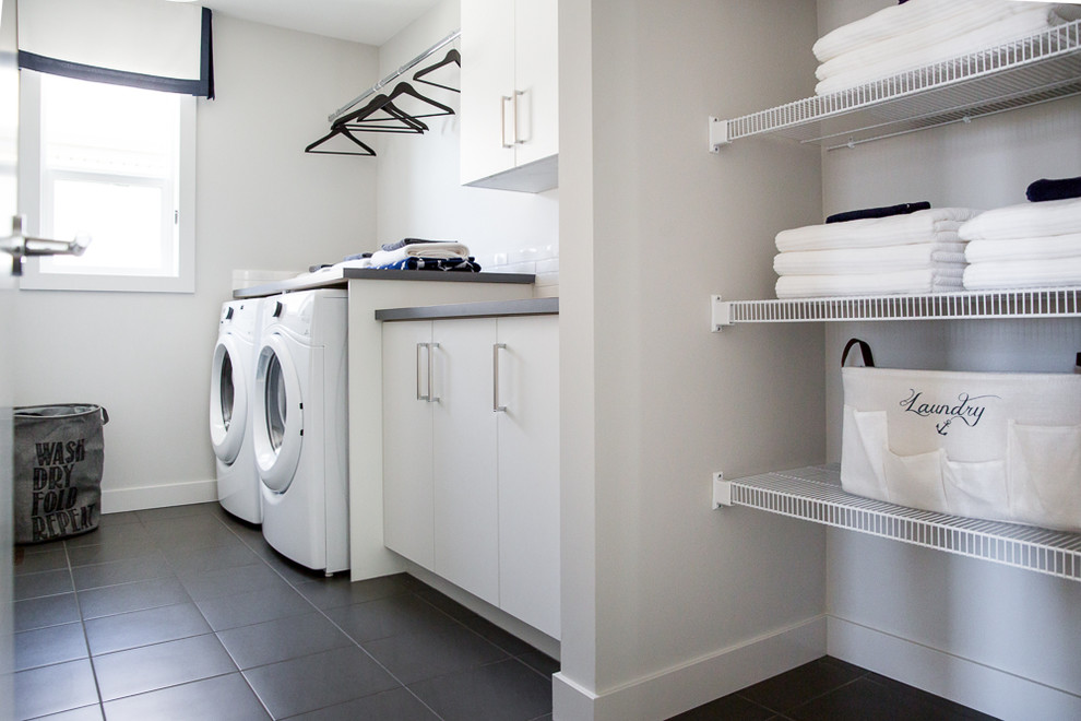 Modern inredning av en mellanstor parallell tvättstuga enbart för tvätt, med släta luckor, vita skåp, bänkskiva i kvarts, vita väggar, klinkergolv i keramik, en tvättmaskin och torktumlare bredvid varandra och grått golv