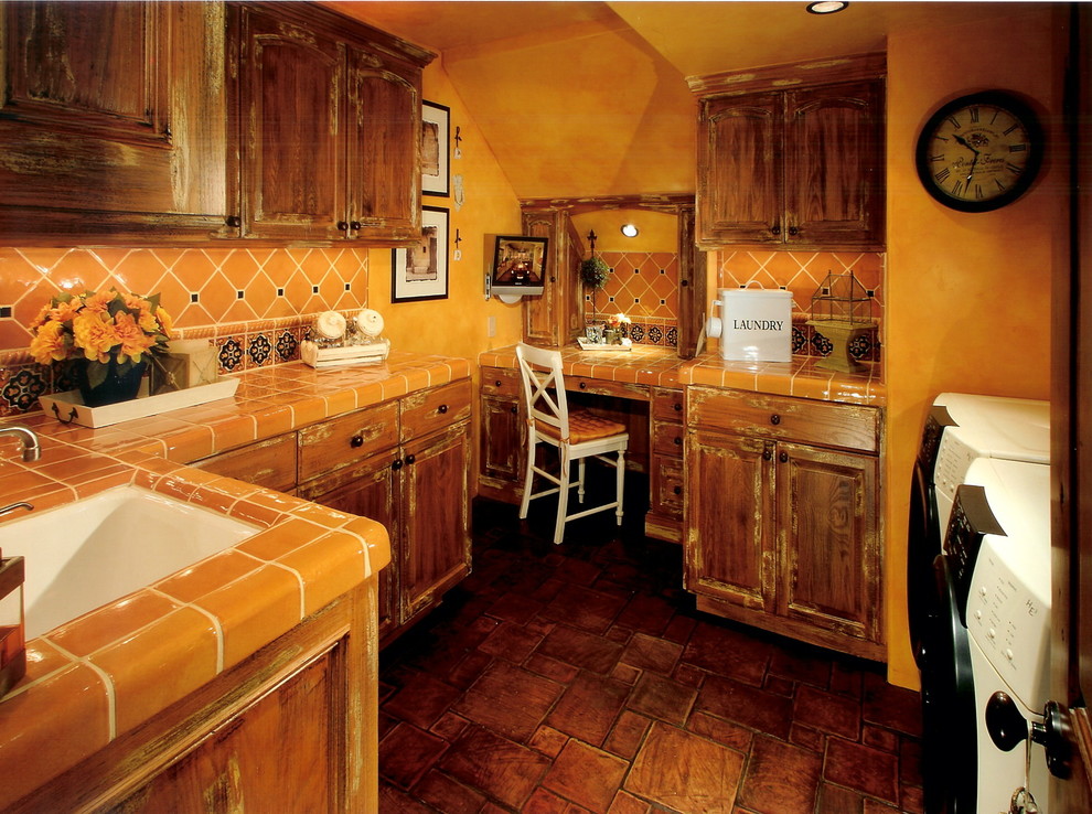 Exempel på en medelhavsstil oranga oranget tvättstuga, med en undermonterad diskho, luckor med upphöjd panel, skåp i slitet trä, kaklad bänkskiva, klinkergolv i terrakotta, en tvättmaskin och torktumlare bredvid varandra och orange väggar
