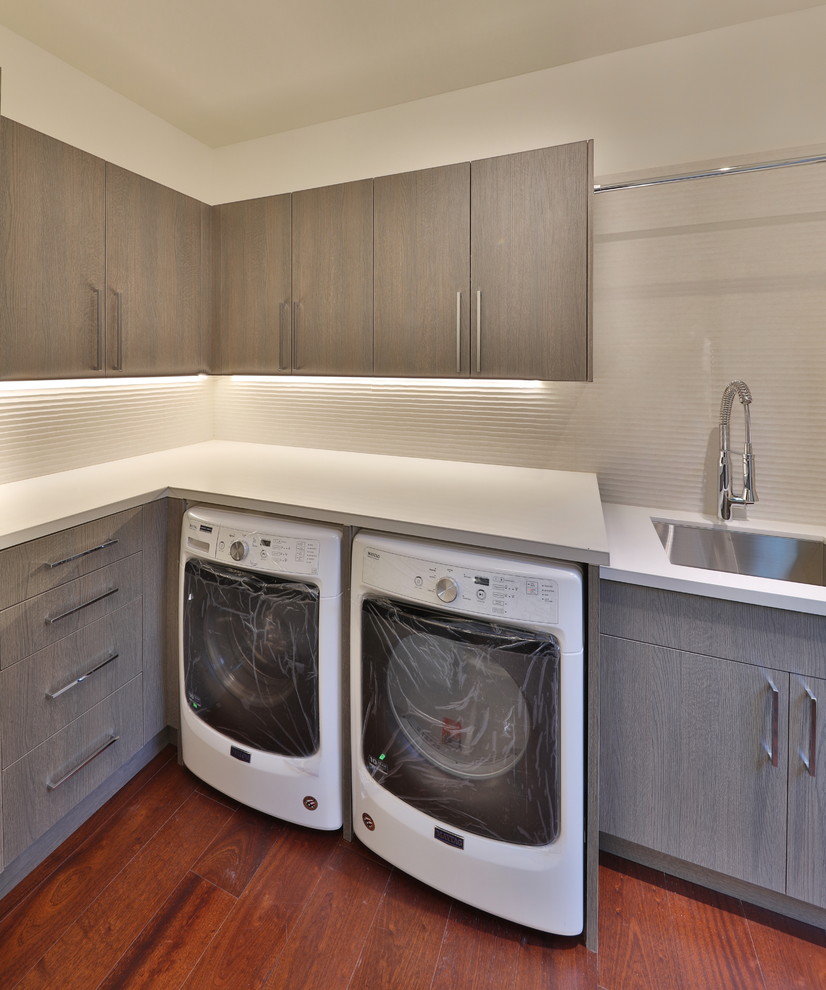 Exempel på en liten modern vita vitt tvättstuga enbart för tvätt, med en nedsänkt diskho, släta luckor, grå skåp, bänkskiva i kvarts, vita väggar, mellanmörkt trägolv, en tvättmaskin och torktumlare bredvid varandra och brunt golv
