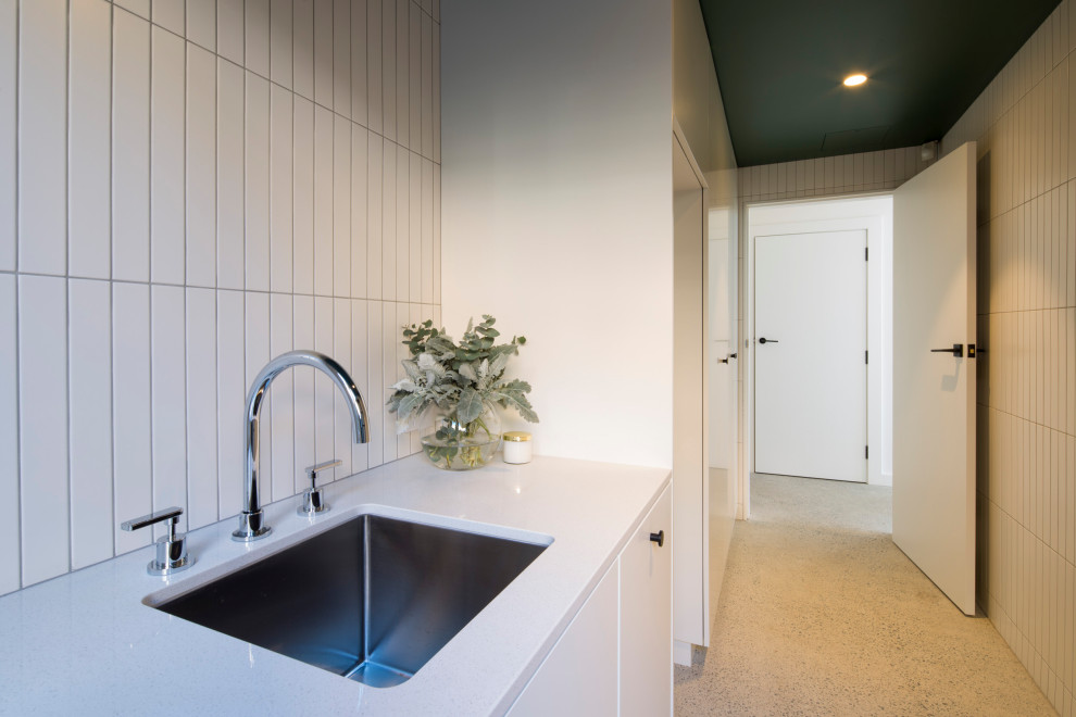 Design ideas for a large contemporary single-wall utility room in Sydney with a single-bowl sink, recessed-panel cabinets, white cabinets, quartz worktops, white walls, concrete flooring, a stacked washer and dryer, grey floors and white worktops.