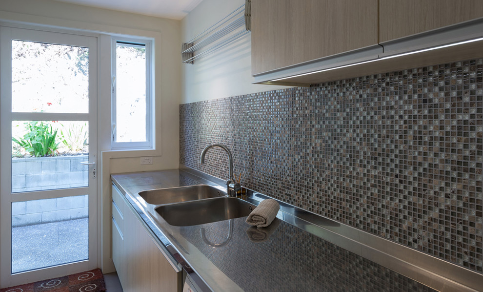 Example of a large minimalist single-wall ceramic tile dedicated laundry room design in Auckland with a double-bowl sink, flat-panel cabinets, stainless steel countertops, white walls, a side-by-side washer/dryer and light wood cabinets