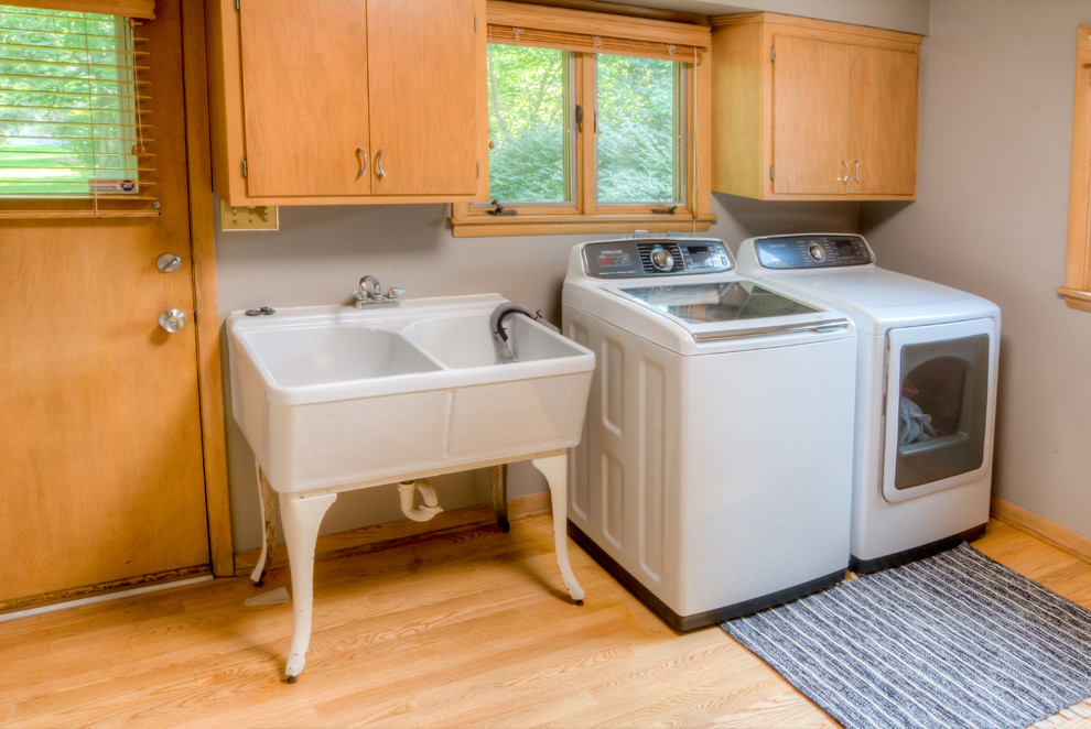 Foto på en mellanstor vintage linjär tvättstuga enbart för tvätt, med en allbänk, släta luckor, skåp i ljust trä, grå väggar, ljust trägolv, en tvättmaskin och torktumlare bredvid varandra och beiget golv