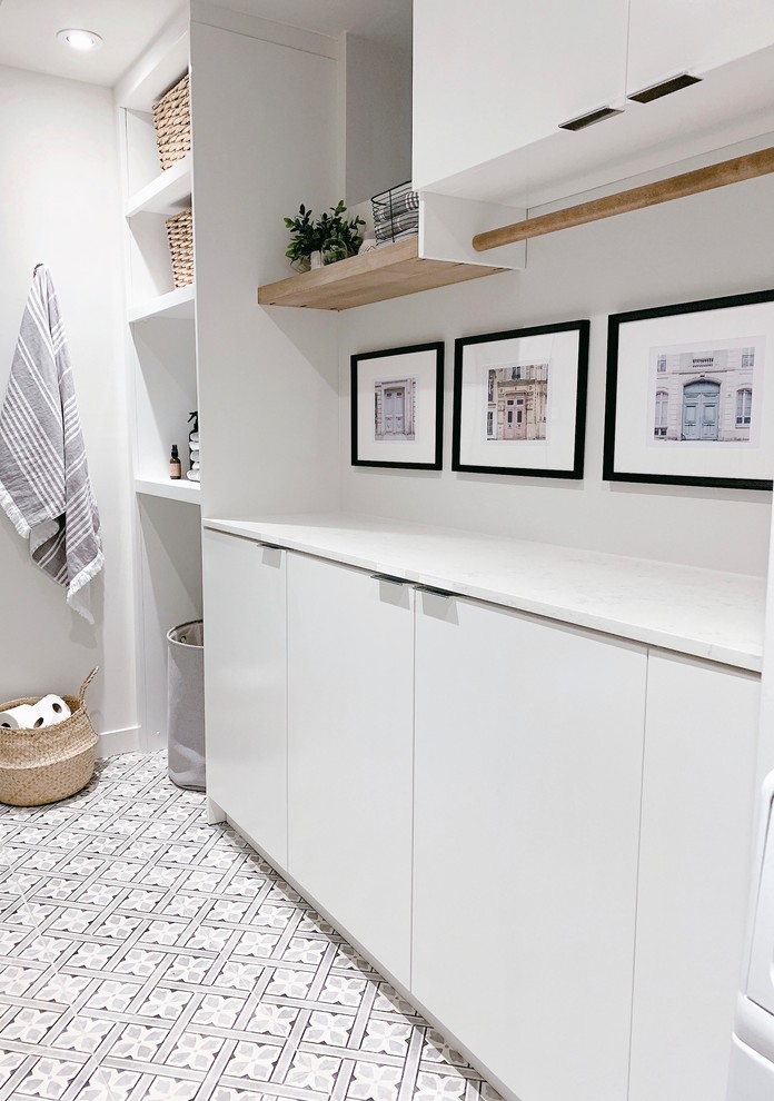 Modern inredning av en vita vitt tvättstuga, med släta luckor, vita skåp, bänkskiva i kvarts, vita väggar, klinkergolv i porslin och en tvättpelare