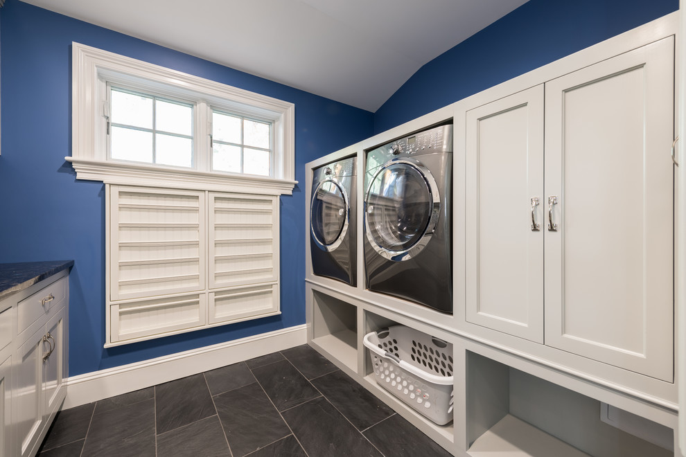 Modelo de cuarto de lavado de galera clásico de tamaño medio con fregadero bajoencimera, puertas de armario blancas, paredes azules, lavadora y secadora juntas, suelo de pizarra y armarios con paneles empotrados