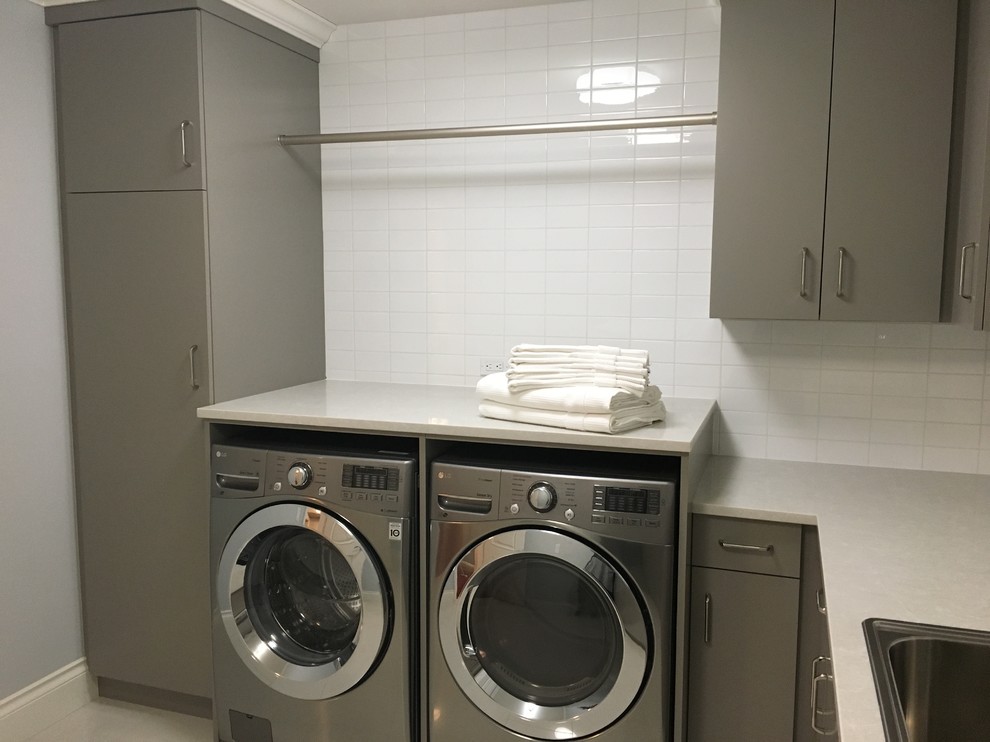 Inspiration for a large transitional l-shaped porcelain tile and white floor dedicated laundry room remodel in Other with a drop-in sink, flat-panel cabinets, gray cabinets, solid surface countertops, gray walls and a side-by-side washer/dryer