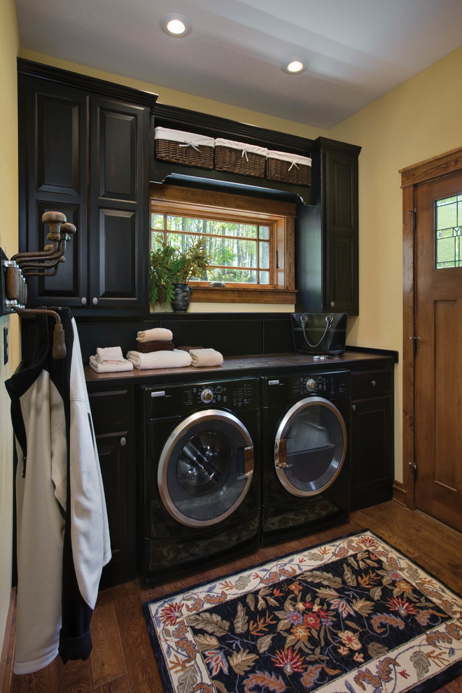 Einzeilige, Mittelgroße Rustikale Waschküche mit schwarzen Schränken, beiger Wandfarbe, dunklem Holzboden, profilierten Schrankfronten und Waschmaschine und Trockner nebeneinander in Sonstige