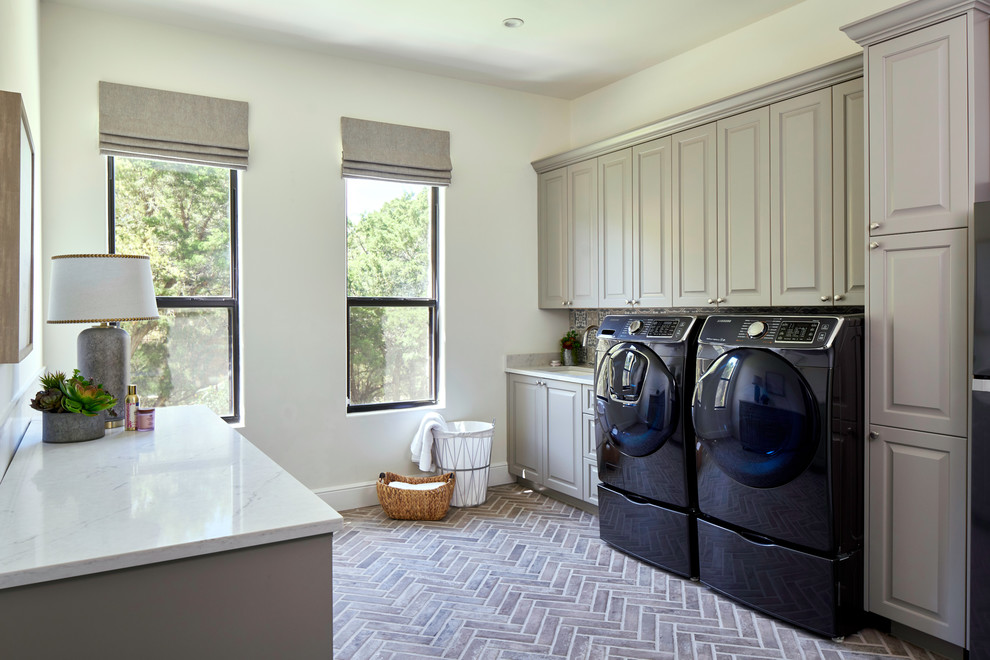 Klassisk inredning av en beige parallell beige tvättstuga, med en undermonterad diskho, luckor med upphöjd panel, grå skåp, vita väggar, tegelgolv, en tvättmaskin och torktumlare bredvid varandra och brunt golv