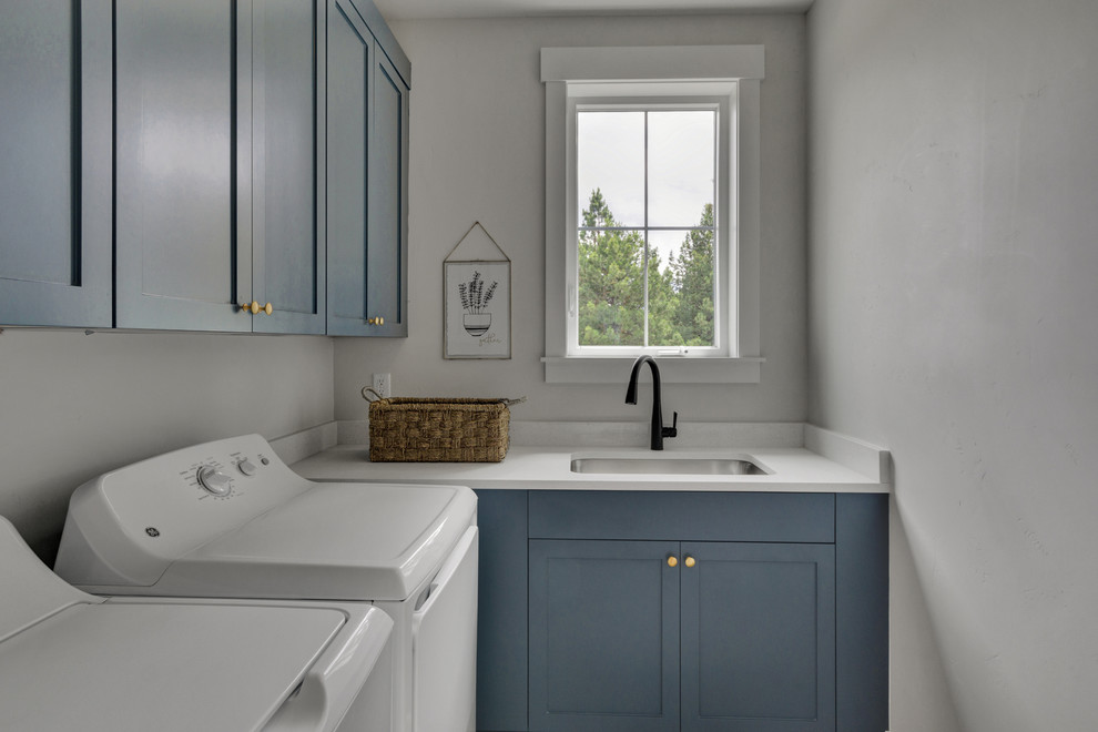 Foto på en liten funkis vita l-formad tvättstuga enbart för tvätt, med en integrerad diskho, luckor med infälld panel, blå skåp, bänkskiva i kvarts, vita väggar, en tvättmaskin och torktumlare bredvid varandra och vitt golv