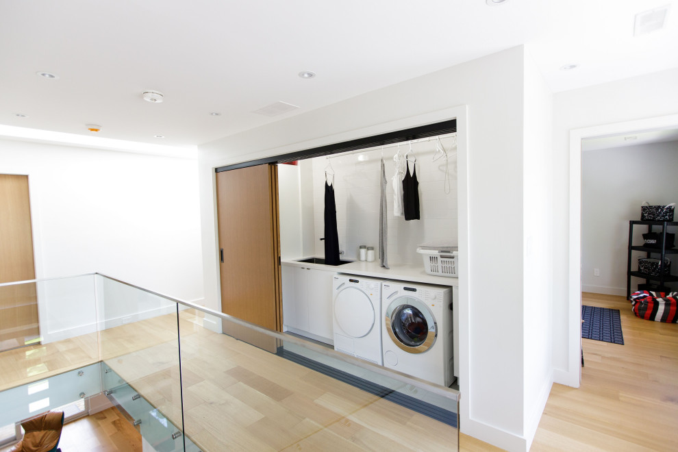 Modern inredning av en vita vitt liten tvättstuga, med en integrerad diskho, öppna hyllor, vita skåp, mellanmörkt trägolv, en tvättmaskin och torktumlare bredvid varandra och brunt golv