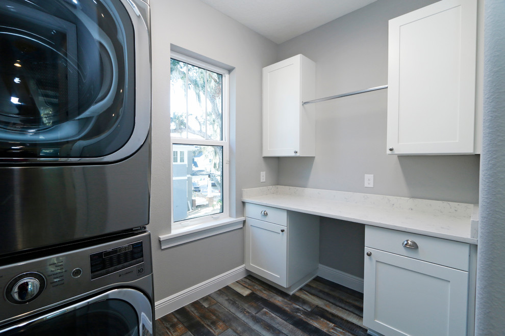 Ejemplo de cuarto de lavado actual de tamaño medio con encimera de cuarzo compacto, paredes grises, suelo de madera oscura, lavadora y secadora apiladas y puertas de armario blancas