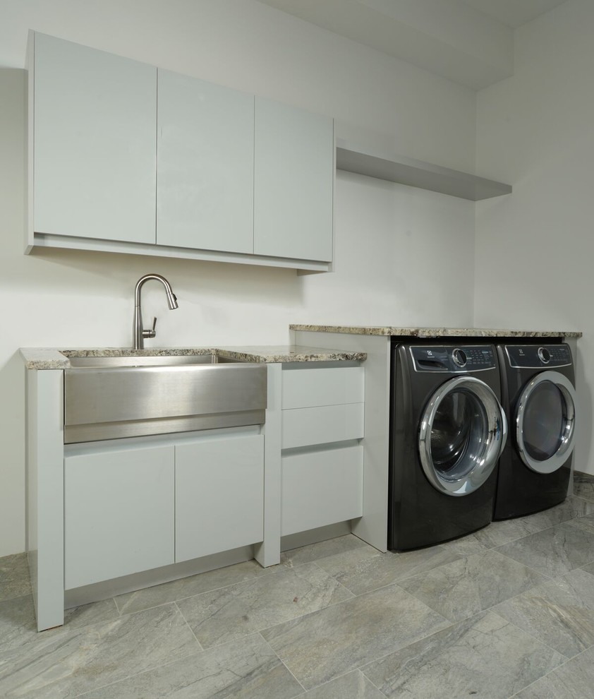 Design ideas for a modern utility room in Orlando with a belfast sink, flat-panel cabinets, white cabinets, granite worktops, white walls, a side by side washer and dryer and grey floors.