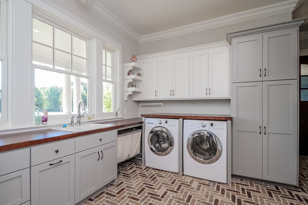 Example of a large transitional l-shaped brown floor dedicated laundry room design in Charlotte with a drop-in sink, shaker cabinets, gray cabinets, wood countertops, gray walls and a side-by-side washer/dryer