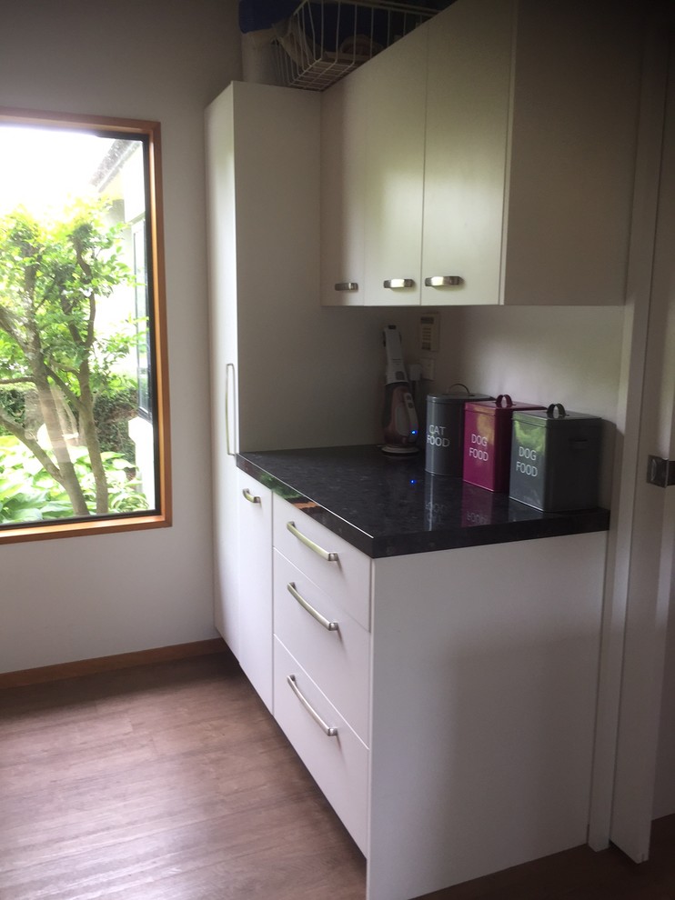 Medium sized contemporary galley separated utility room in Christchurch with a single-bowl sink, flat-panel cabinets, white cabinets, laminate countertops, beige walls, vinyl flooring and a stacked washer and dryer.
