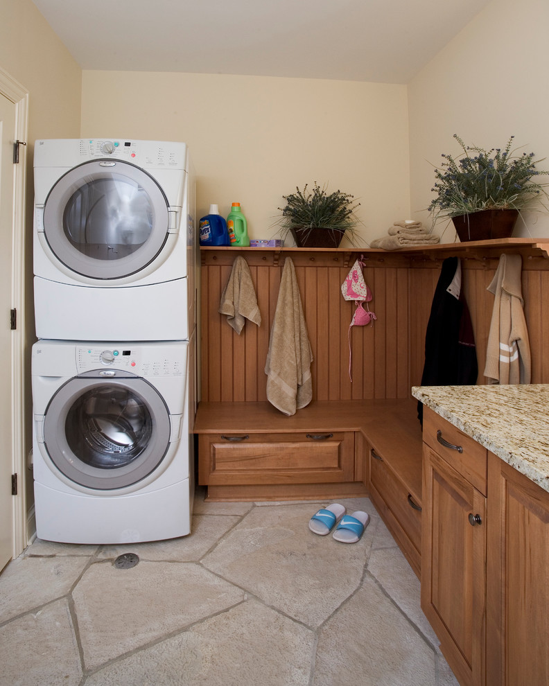 Example of a large classic l-shaped limestone floor dedicated laundry room design in Chicago with an undermount sink, raised-panel cabinets, medium tone wood cabinets, granite countertops, beige walls and a stacked washer/dryer