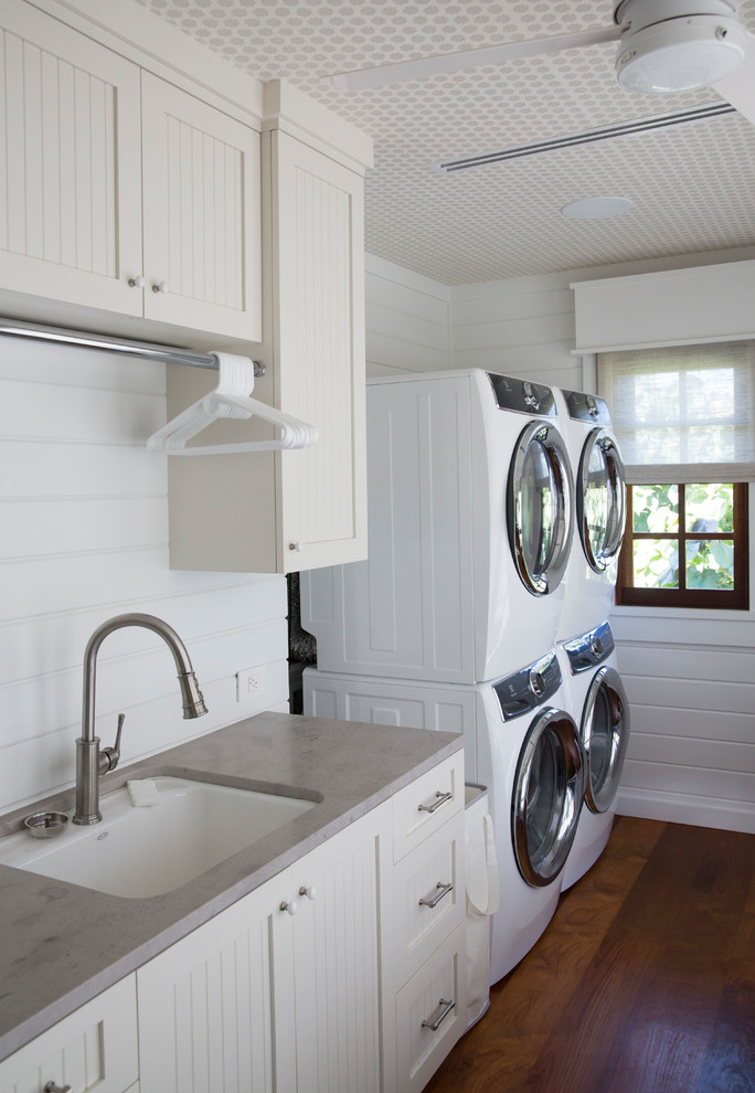 Inspiration for a tropical single-wall medium tone wood floor and brown floor dedicated laundry room remodel in Hawaii with a farmhouse sink, louvered cabinets, white cabinets, white walls, a stacked washer/dryer and gray countertops