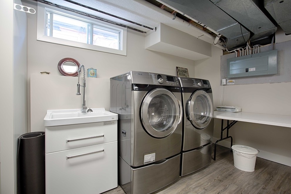 Diseño de cuarto de lavado lineal urbano de tamaño medio con pila para lavar, armarios con paneles lisos, puertas de armario blancas, paredes blancas, suelo de madera oscura y lavadora y secadora juntas