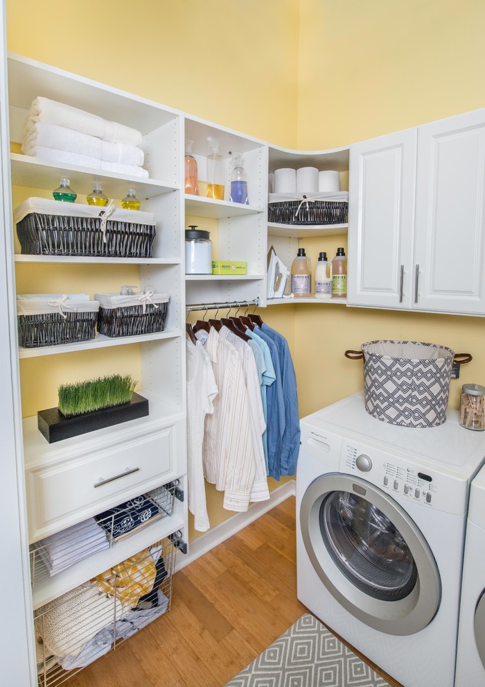 Imagen de lavadero en L de tamaño medio con armarios con paneles con relieve, puertas de armario blancas, paredes amarillas, suelo de bambú y lavadora y secadora juntas