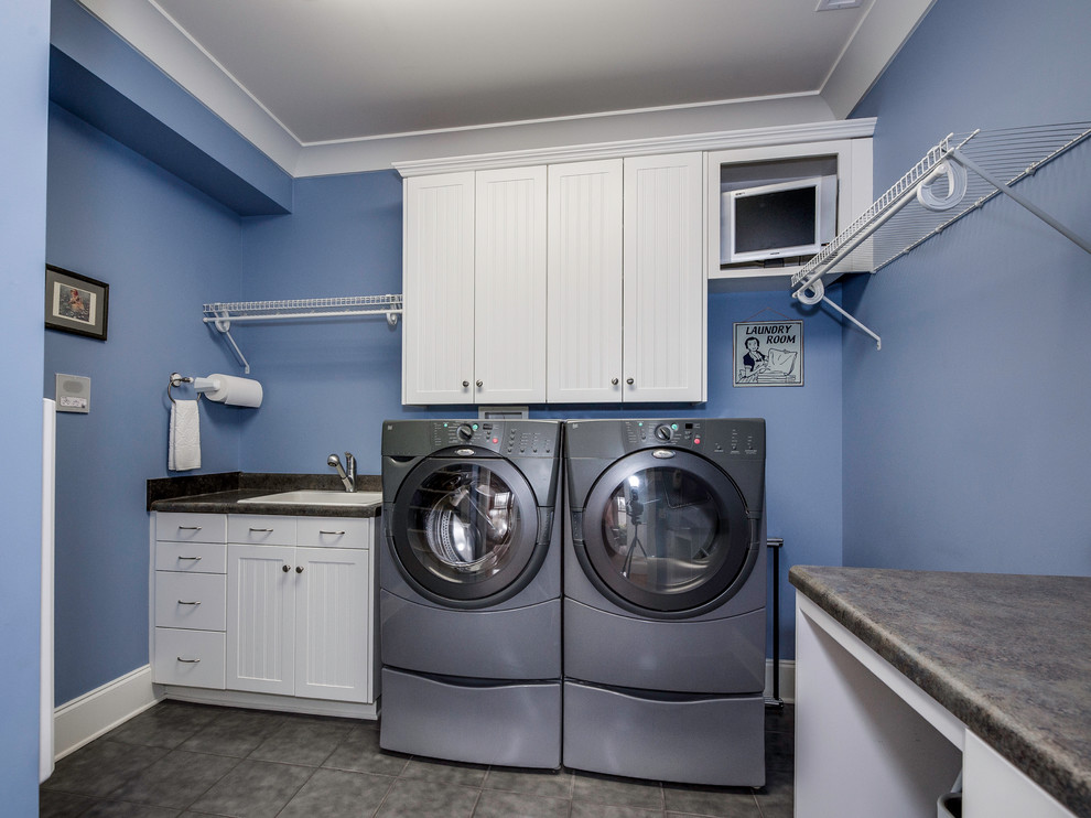 Modelo de cuarto de lavado de estilo americano grande con fregadero de un seno, armarios con rebordes decorativos, puertas de armario blancas, encimera de laminado, paredes azules, suelo de baldosas de porcelana y lavadora y secadora juntas