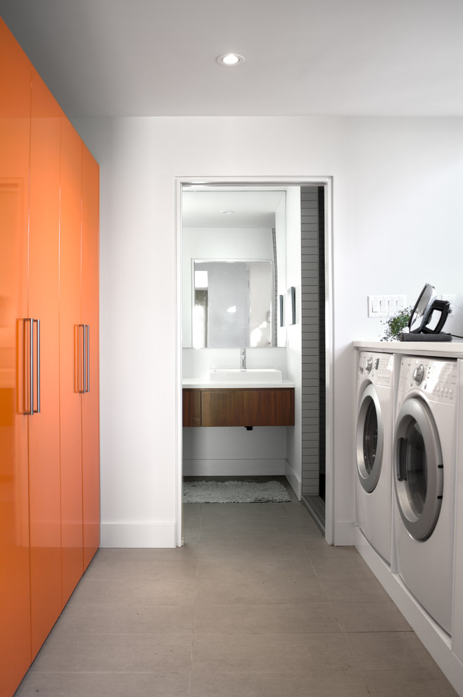 他の地域にあるミッドセンチュリースタイルのおしゃれな洗濯室 (ll型、フラットパネル扉のキャビネット、オレンジのキャビネット、白い壁、左右配置の洗濯機・乾燥機、白いキッチンカウンター) の写真