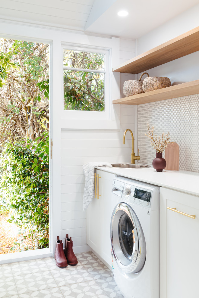 Lantlig inredning av en tvättstuga, med en nedsänkt diskho, vita väggar, en tvättmaskin och torktumlare bredvid varandra och grått golv